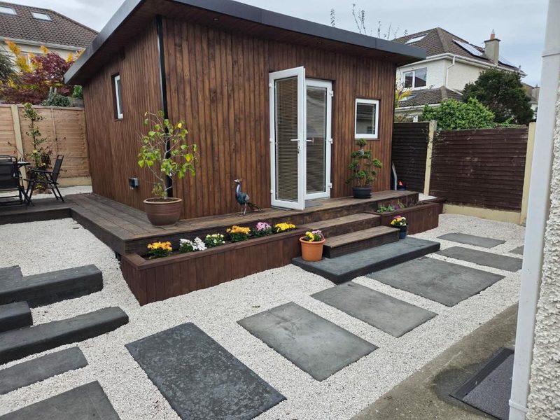 A timber clad garden room with French doors, a raised deck with steps and built in planters, set in a gravelled garden