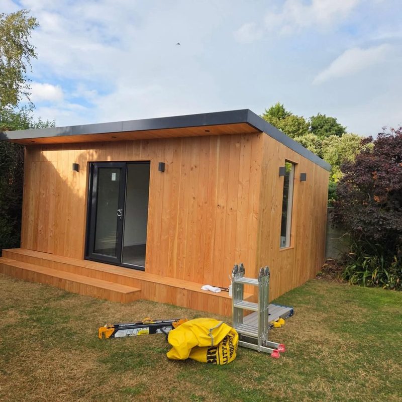 A garden room clad in natural timber with a flat roof, anthracite sliding patio doors and a timber deck running the full width of the front