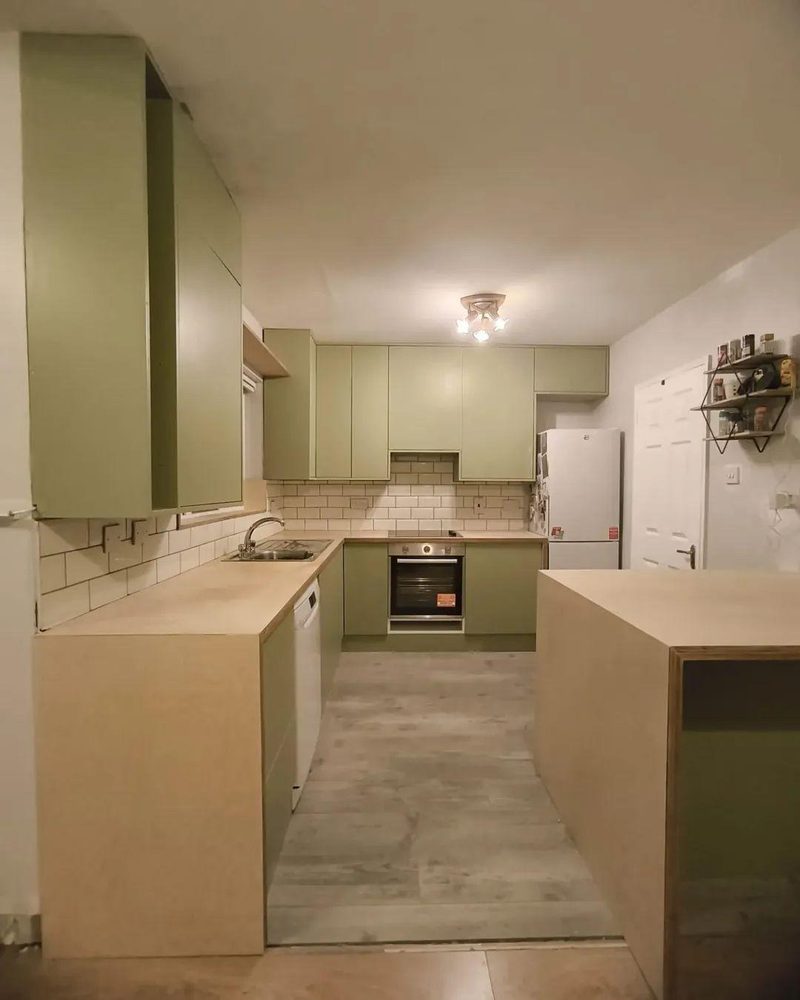 A fitted kitchen with sage green doors, birch plywood worktops and a white metro tile splashback, seen from the doorway