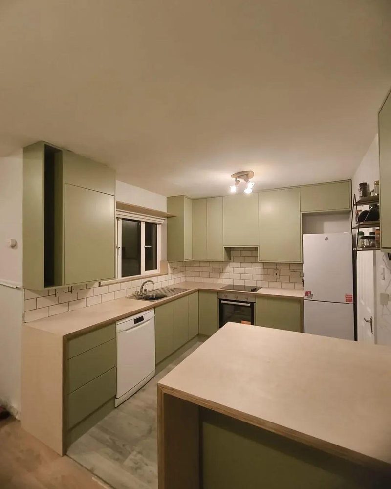 A sage green fitted kitchen with an integrated oven and hob, dishwasher and a run of birch plywood worktop under a window