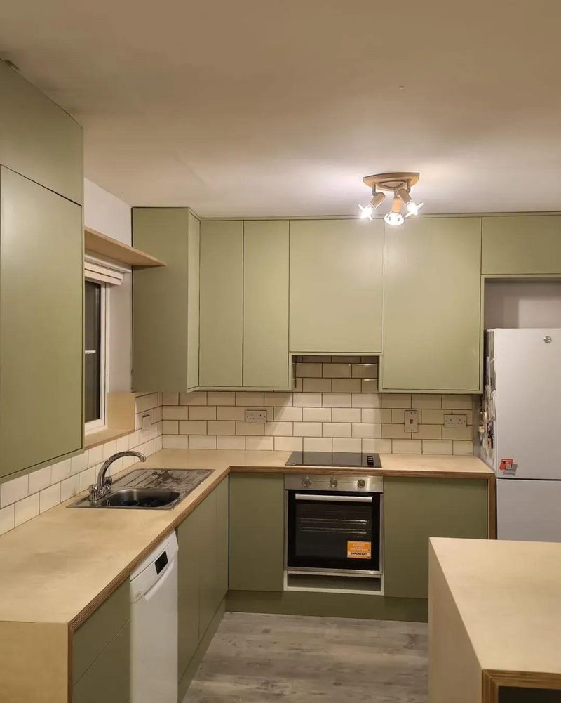 A sage green fitted kitchen showing the sink, dishwasher, induction hob and built in oven under birch plywood worktops